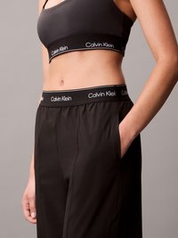 Calças de treino pretas com uma cintura larga, apresentando um logótipo branco. Tecido suave com bolsos laterais e um corte descontraído.