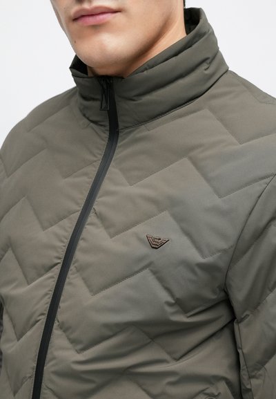 Prešívaná bunda v olivovo zelenej farbe s vysokým golierom, vybavená čiernym zipsom a hnedým logom na ľavej strane.