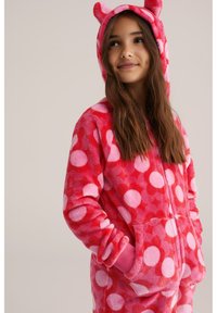 Švelnus rožinis fliso kombinezonas su didelėmis įvairiaspalvėmis taškelėmis rožinės spalvos atspalviuose, su kapučinu, meškos ausimis ir priekinėmis kišenėmis. Minkšta tekstūra.