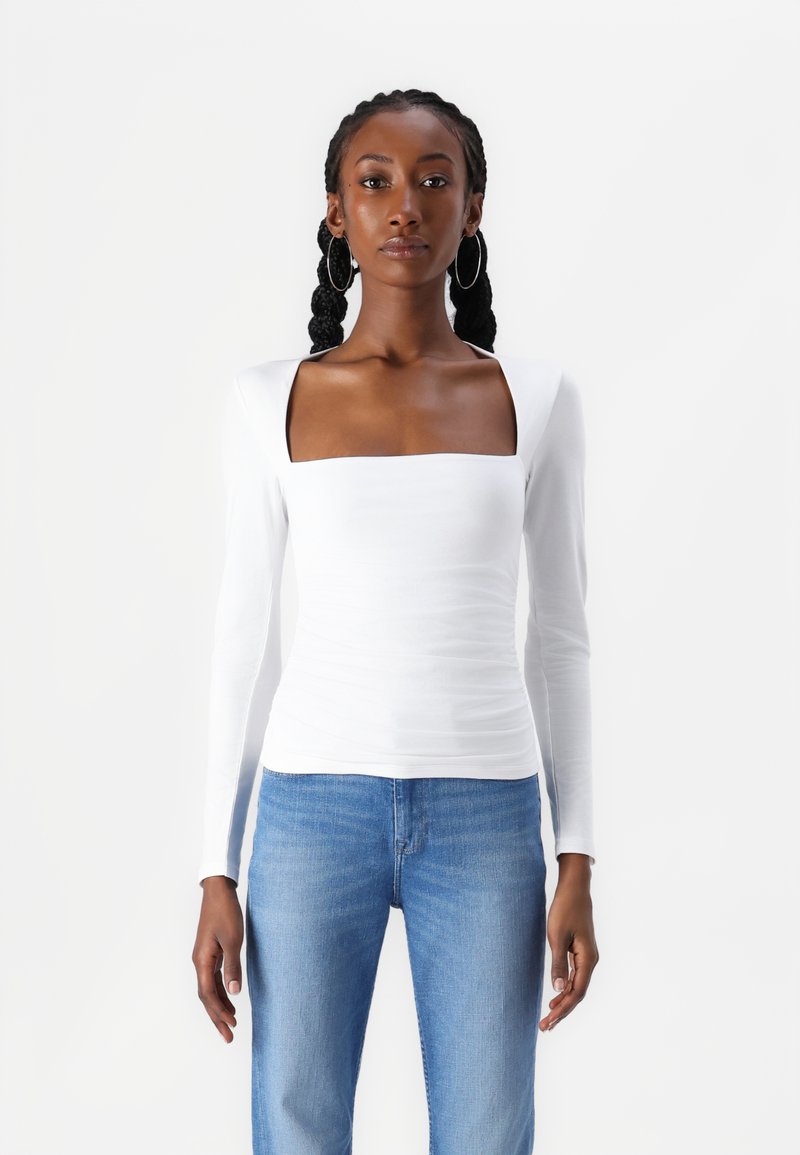 Long-sleeve white top with a square neckline and fitted design, paired with light blue jeans. Smooth fabric with subtle ruching details.