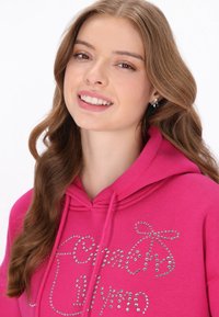 Sudadera rosa con capucha y adornos de piedras de strass en el frente. La modelo tiene el cabello largo, ondulado y castaño, y está sonriendo.