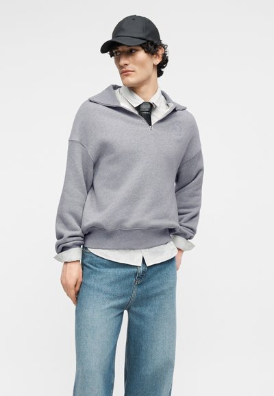 Νεαρός άνδρας που φοράει μαύρο καπέλο, γκρι φούτερ με φερμουάρ μέχρι το στήθος πάνω από ριγέ πουκάμισο και μαύρη γραβάτα, με μπλε τζιν, τα χέρια στις τσέπες.