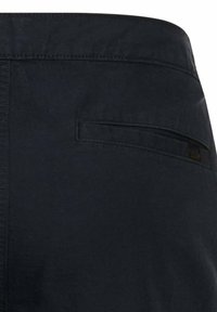Gros plan sur un pantalon en tissu noir montrant une poche passepoilée horizontale à l'arrière avec des détails de couture subtils.