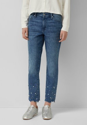 Persona che indossa jeans di denim blu con orli sfrangiati e piccoli dettagli di perle, abbinati a sneaker argentate e una maglia bianca a maniche lunghe.