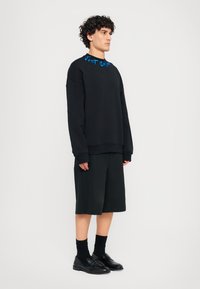 Sweatshirt preto com texto azul na gola, combinado com calções pretos até aos joelhos e sapatos pretos. Tecido suave, ajustamento relaxado.