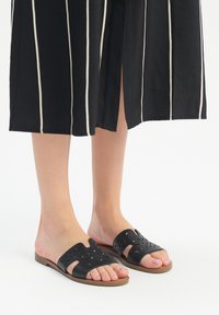 Czarne skórzane sandały wsuwane z wycięciami, płaską podeszwą i jasnobrązową podeszwą, noszone w połączeniu z sukienką w czarno-białe paski.