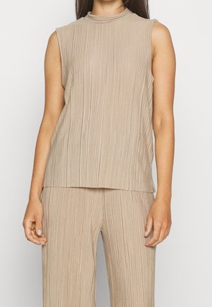 Kvinde iført beige ærmeløs ribstrikket top og matchende ribstrikkede bukser mod en ensfarvet hvid baggrund.