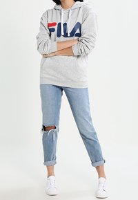 Persona que lleva una sudadera gris de FILA, jeans claros rasgados enrollados en los tobillos y zapatillas blancas, de pie con los brazos cruzados.