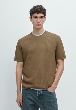 Jonge man die een bruin textuur kortarmtrui draagt over een wit overhemd en blauwe spijkerbroek, staand met één hand in de zak.