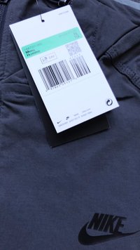 Pilkas „Nike“ marškinėliai su juodu logotipu, kuriuos lydi etiketė su dydžiu XL, brūkšniniu kodu ir prekės ženklo informacija. Lygus audinys su subtiliu žvilgesiu.
