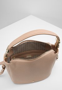 Béžová kožená kabelka s texturou, širokým popruhem, zlatými kovovými prvky, zapínáním na zip a vzorovanou látkovou podšívkou, která ukazuje malou kapsu na zip.