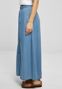 Denim maxi skjørt med elastisk belte, knappedetaljer på siden, og en lys blå farge. Kombinert med brune vevde sandaler.