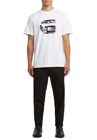 Hombre con camiseta blanca de manga corta con un diseño gráfico abstracto negro, pantalones negros y zapatos negros, de pie recto frente a un fondo blanco.