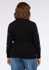 Svart cardigan med långa ärmar, knytmidja och silverfärgade rosettdekor på ärmarna, tillverkad av ett mjukt stickat material.