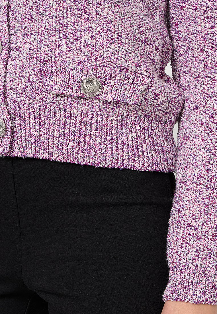 Texturiertes pinkes und lila Cardigan mit geripptem Saum und Ärmeln, ausgestattet mit zwei silbernen Knöpfen zum Verschließen. Darunter werden schwarze Hosen getragen.