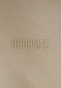 Beige tekstil med en struktureret overflade med ordet "DROPSIZE" prægede i en matchende farve.
