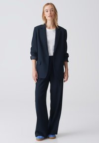 Costume bleu marine avec une veste cintrée et un pantalon large. La veste a des manches retroussées, associée à un t-shirt blanc et des chaussures bleues.