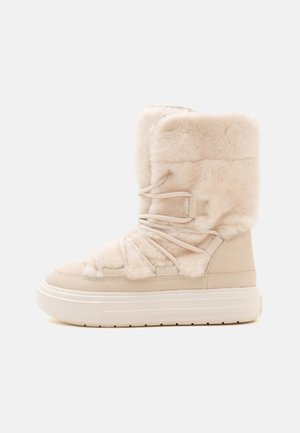 Botte d’hiver beige clair avec une semelle épaisse en caoutchouc blanc, une tige en peluche douce et des lacets décoratifs entourants.