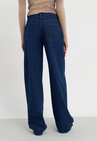 Henkilö, joka käyttää korkeavyötäröisiä, leveälahkeisia tummansiniä farkkuja ja vaaleanbeigeä ribattyä pitkähihaista paitaa, seisoo yksinkertaista valkoista seinää vasten.