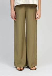 Pantalons larges en olive, fabriqués dans un tissu léger, avec une texture lisse et des coutures discrètes, associés à des tongs marron.