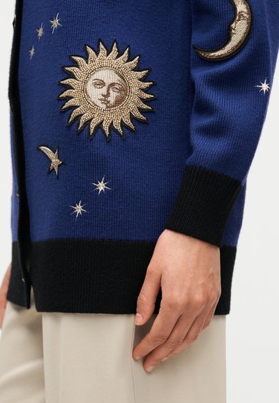 Cardigan bleu tricoté avec broderies soleil et lune, accents en fil d'or, bordures noires et texture lisse. Détail de motifs d'étoiles.