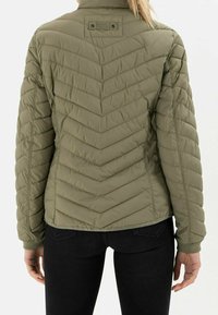 Frau in einer olivgrünen gesteppten Jacke mit Chevron-Steppung, steht mit dem Rücken zur Kamera, kombiniert mit schwarzen Hosen.