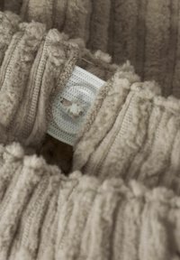 Bežinis plush audinys su vertikalių briaunų raštu ir minkštais pom-pom akcentais, su balta etikete ir apvaliu mygtuko detaliu.
