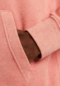 Różowa bluza z kapturem w plamy, z przednią kieszenią; widoczna ręka w kieszeni, pokazująca teksturowany ciemno brązowy odcień skóry. Miękka tkanina i detal ściągacza.