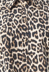 Leopardenmusterhemd mit beigem Grund und schwarzen sowie braunen Flecken, Knopfschluss und Kragen. Glatte Textur.