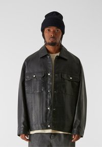 Schwarzes Leder-Übershirt mit lockerer Passform, zwei vorderen Taschen, silbernen Druckknöpfen und einer gerippten schwarzen Mütze. Der Stoff hat einen leichten Glanz.