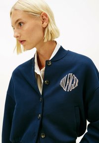 Mujer rubia con chaqueta azul marino abotonada con el logo "HILFIGER" sobre una camisa blanca con cuello, mirando hacia la izquierda contra un fondo liso.