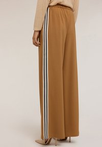 Bruine wijde broek met een elastische tailleband, voorzien van verticale driestrepen in zwart, wit en grijs aan de zijkanten.