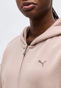 Κοντινό πλάνο ενός ατόμου που φοράει ένα φούτερ με φερμουάρ Puma σε ανοιχτό ροζ χρώμα με το λογότυπο της Puma στο στήθος, μπροστά σε απλό φόντο.