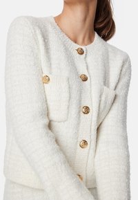 Vit texturerad cardigan med rund halsringning, med guldfärgade knappar och en framficka. Mjuk tyg med en mysig känsla.