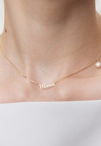 Złoty naszyjnik z zawieszką "Mama", z delikatnym łańcuszkiem i małym perłowym akcentem, na tle nagiej skóry.