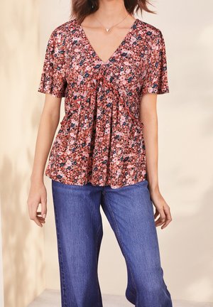 Blusa floral en tonos rosa y azul marino con cuello en V, mangas cortas con vuelo y detalles fruncidos. Combinada con vaqueros de pierna ancha en azul claro.