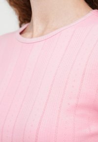 Κοντινό πλάνο ενός ατόμου που φοράει ανοιχτό ροζ υφασμένο τοπ με κάθετες ριγέ μοτίβο και στρογγυλή λαιμόκοψη.