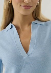 Žena v svetlom modrom pletenom topi s hlbokým V-výstrihom a malým golierikom, doplnená okrúhlymi zlatými náušnicami.