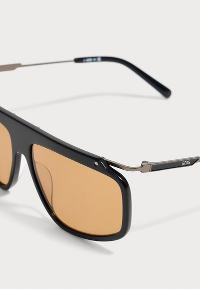Lunettes de soleil rectangulaires noires avec des verres teintés marron et des branches métalliques fines, avec le logo GCDS à l’extrémité des branches.