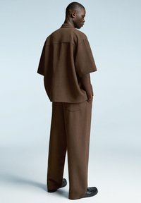 Rjava, ohlapna kratka majica z kratkimi rokavi in široki hlačami, ki imajo teksturirano tkanino. Maneken nosi črne natikače.