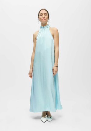 Femme portant une robe maxi sans manches bleu clair à col halter et des chaussures blanches pointues, debout sur un fond blanc uni.