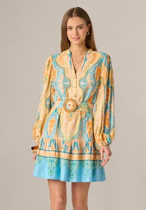 Jeune femme portant une robe à manches longues avec des motifs, et une grande boucle de ceinture ronde tressée, debout devant un fond uni.