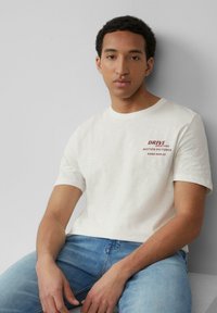 Witte katoenen T-shirt met korte mouwen, voorzien van rode opdruk "DRIVE" en "MOTION PICTURES." Gepaard met lichtblauwe denim jeans.