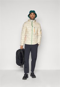 Beige puffer jacket with quilted design and multiple zip pockets. Dark pants, black backpack, and green bucket hat. Black sneakers.