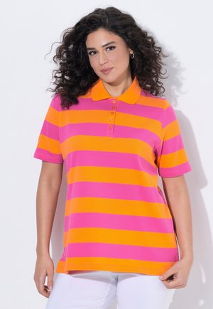 Femme aux cheveux bouclés et foncés portant un polo à manches courtes avec des rayures horizontales orange et rose, associé à un pantalon blanc.