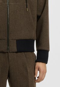 Bruin herringbone zip-up jack met geribbelde marineblauwe manchetten. Voorzien van voorzakken en een capuchon met trekkoord; gemaakt van een textuurwol blend.