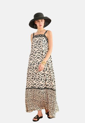 Mujer con un vestido maxi beige con patrones de hojas negras, sombrero negro de ala ancha, joyas doradas y sandalias negras, de pie frente a un fondo liso.