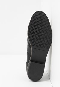 Semelle de chaussure noire avec motif à crampons en grille et indication de taille 37, vue par le dessous sur fond blanc.