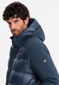 Marineblaue Steppjacke mit abnehmbarer Kapuze, die eine Kombination aus matten und glänzenden Texturen, Druckknöpfen und Taschendetails aufweist.