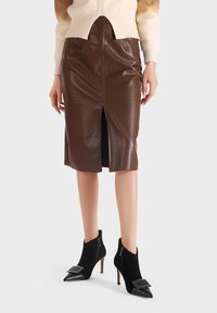 Gonna a tubino in finta pelle marrone con spacco frontale, abbinata a stivaletti neri alla caviglia con punte appuntite e fibbie quadrate.